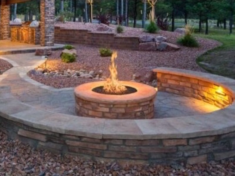 Gas fire pit with flame set into a stone hardscape patio at dusk